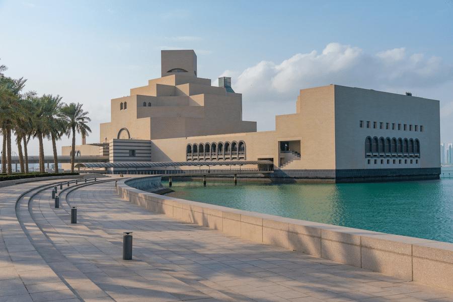 The Museum of Islamic Art in Doha, Qatar stands impressively by the turquoise waters. Palm trees line the curved walkway in the foreground. A visit is a must in Qatar.