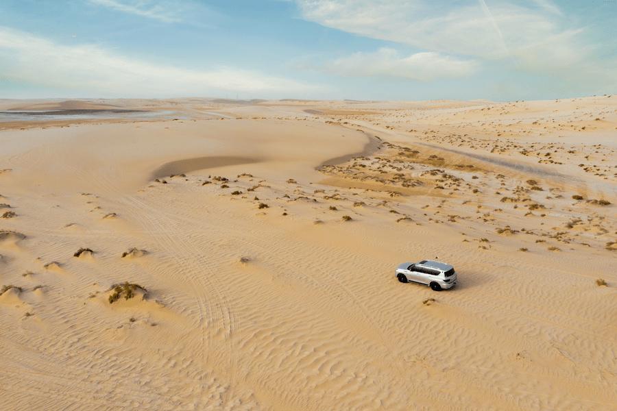 A white SUV drives through the vast yellow-brown sand desert of Qatar. The blue sky is dotted with light clouds. The desert is ideal for an adventurous desert safari.