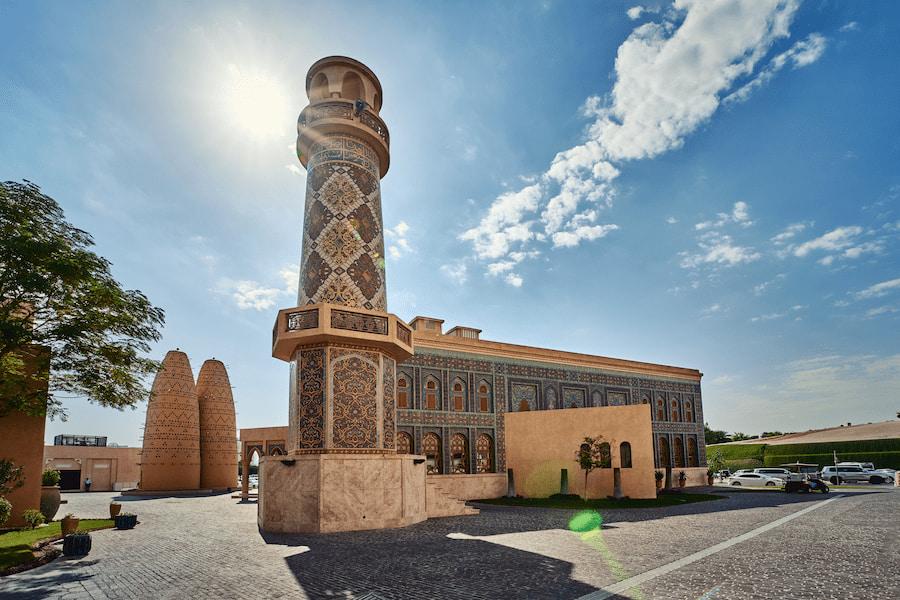 An elaborately decorated minaret rises into the sun-blue sky, flanked by two traditional pigeon towers to the left. To the right, a richly decorated building extends. Cars are parked along the edge.