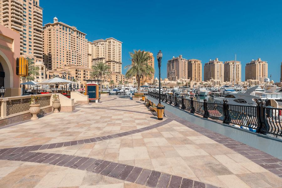 Promenade in Marina Bay, Qatar, with luxurious yachts on the right bank. In the background are tall beige skyscrapers. Palm trees and seating areas under umbrellas create a relaxed atmosphere.