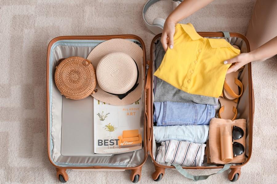 An open brown suitcase lies on a beige carpet. Travel items such as clothing, sunglasses, and a sun hat are already inside. A person is adding another yellow shirt.
