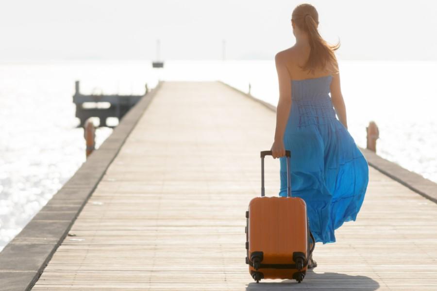 A woman in a blue dress pulls an orange suitcase behind her as she walks over a pier.
