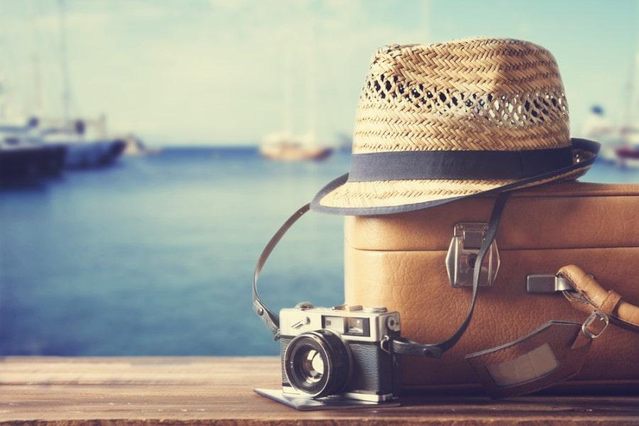 In the foreground on the right is a brown suitcase, in front a camera, and on it a hat. In the background is a harbor, with blue water and ships.