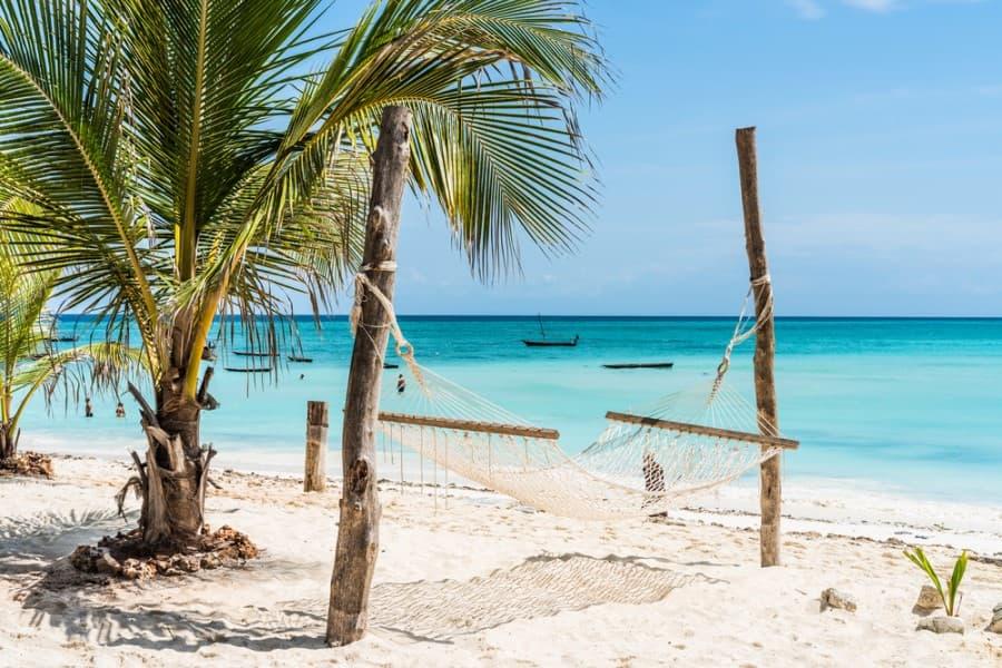 Fine white sand beach with a hammock between two palm trees. The sea is turquoise and calm, a wooden boat rests on the water in the background. Perfect for a relaxing vacation in Zanzibar.