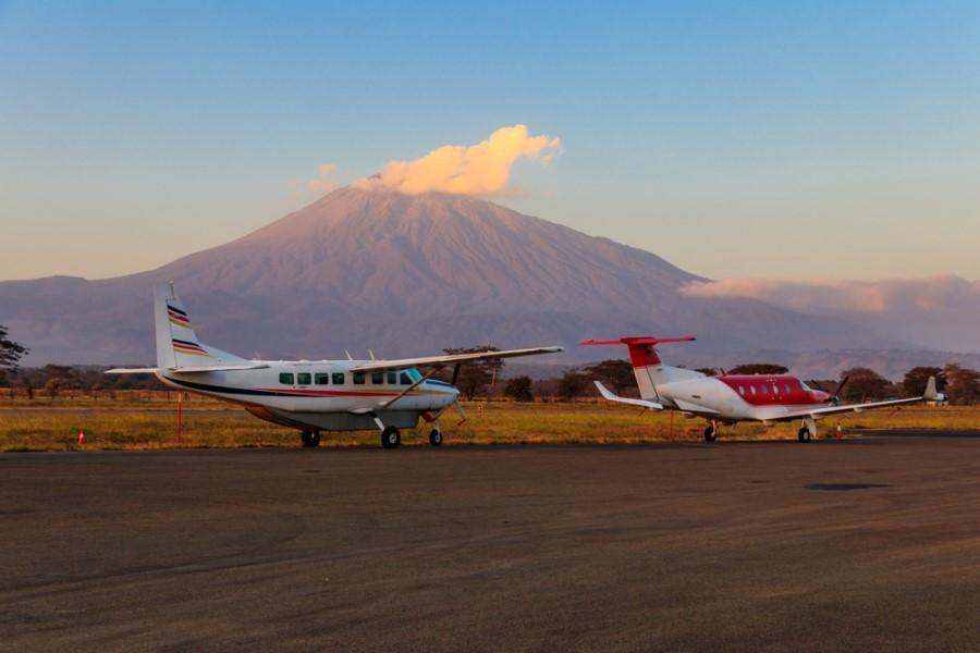 Small planes on a runway at sunset. In the background, a majestic mountain rises. In the foreground, grassy plains are visible, and the sky shines softly.