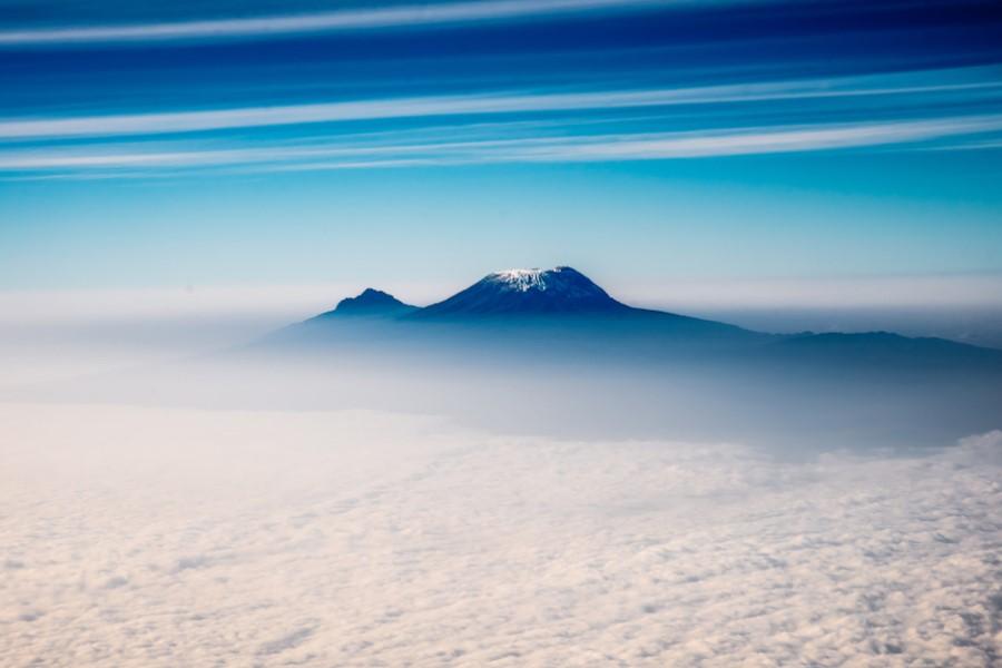 Freestanding snow-covered mountain Kilimanjaro in the center of the image, under a clear blue sky with scattered clouds. A dense cloud layer covers the lower part of the image.