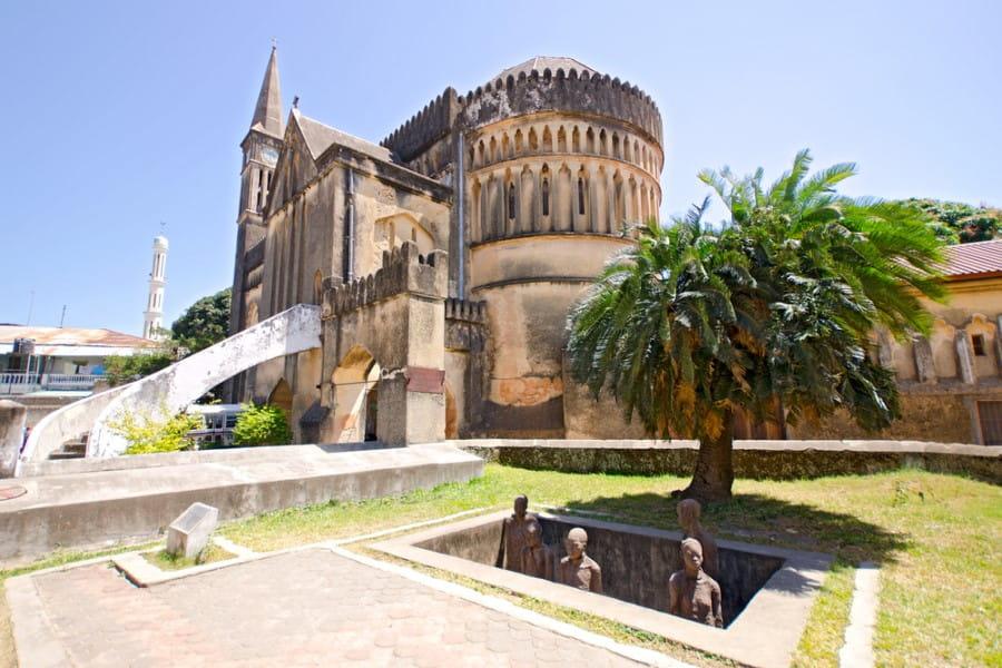 Tall cathedral in the background with a massive tower and large dome, surrounded by palm trees and green plants. In the foreground, there is a fountain and statues. Bright blue sky above.