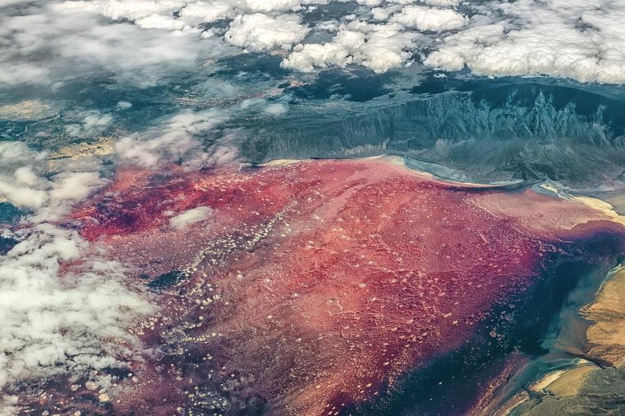 A colorful lake with deep red and violet water, surrounded by cloudy, mountainous landscapes in the background and a few white clouds in the foreground. The colors of the water softly blend together.