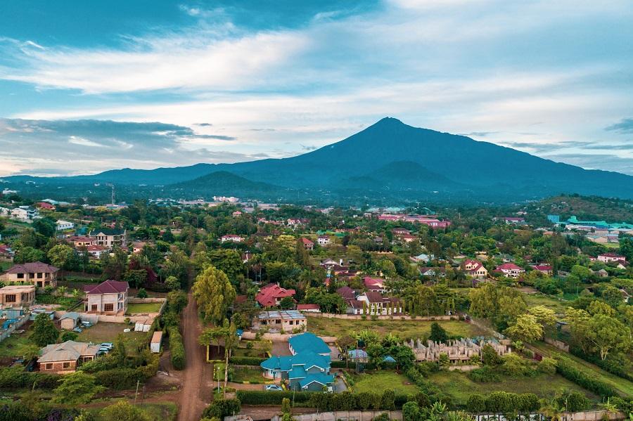 View of Arusha in Tanzania with colorful houses, lots of greenery, and forested hills in the foreground. Majestic Mount Meru rises in the background, surrounded by clouds.