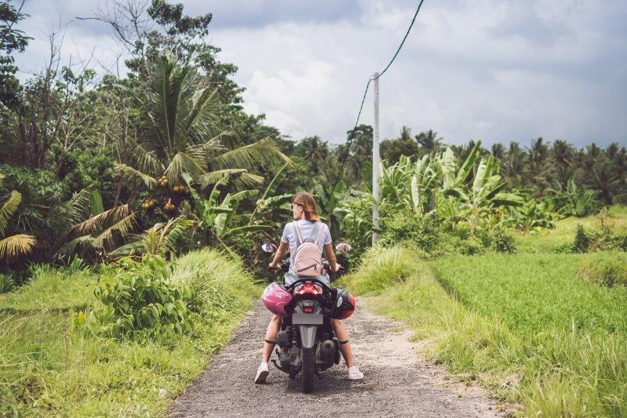 A woman sits on a scooter in Bali on an unpaved road. In the background, you see dense forest.