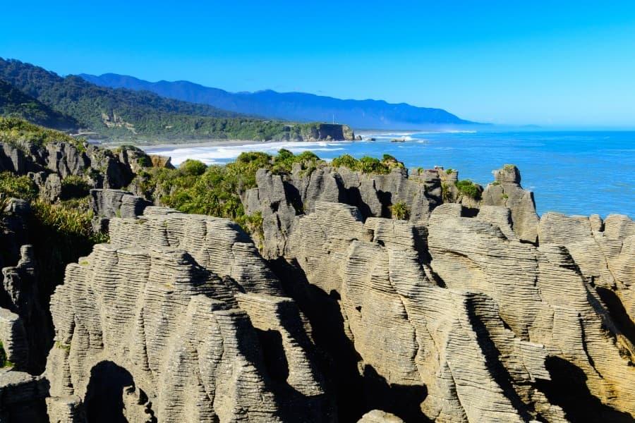 In the foreground of this stunning sight in New Zealand's South Island, the strikingly layered Pancake Rocks of Punakaiki resemble stacks of pancakes, while in the background the deep blue sky and calm sea create a soothing contrast.