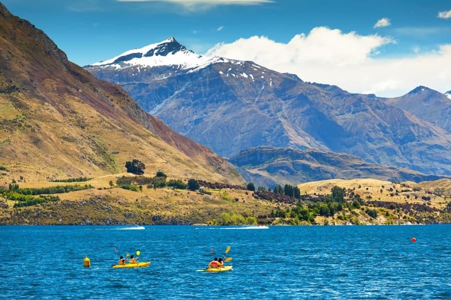On the vibrant photo, you can see kayakers on the clear, blue waters of Lake Wanaka, surrounded by golden hills and towering snow-capped mountains in the background, reflecting the breathtaking beauty of New Zealand's South Island.