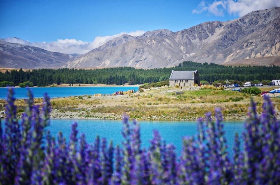 A picturesque view of the "Church of the Good Shepherd" on the shore of turquoise Lake Tekapo, surrounded by blooming lupins in the foreground and majestic mountains in the background, creating a sense of tranquility and beauty typical of the sights of New Zealand's South Island.