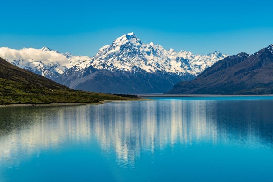 The iconic Mount Cook majestically reflects in the calm, clear waters of Lake Pukaki on New Zealand's South Island, framed by snow-covered peaks and a sky of deep blue, conveying a sense of tranquility and grandeur.