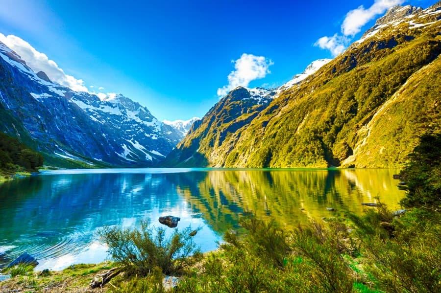 A picturesque depiction of Fiordland National Park, one of the sights on New Zealand's South Island, with a clear, calm lake in the foreground, surrounded by majestic snow-capped mountains and lush green slopes, conveying a sense of grandeur and unspoiled nature.