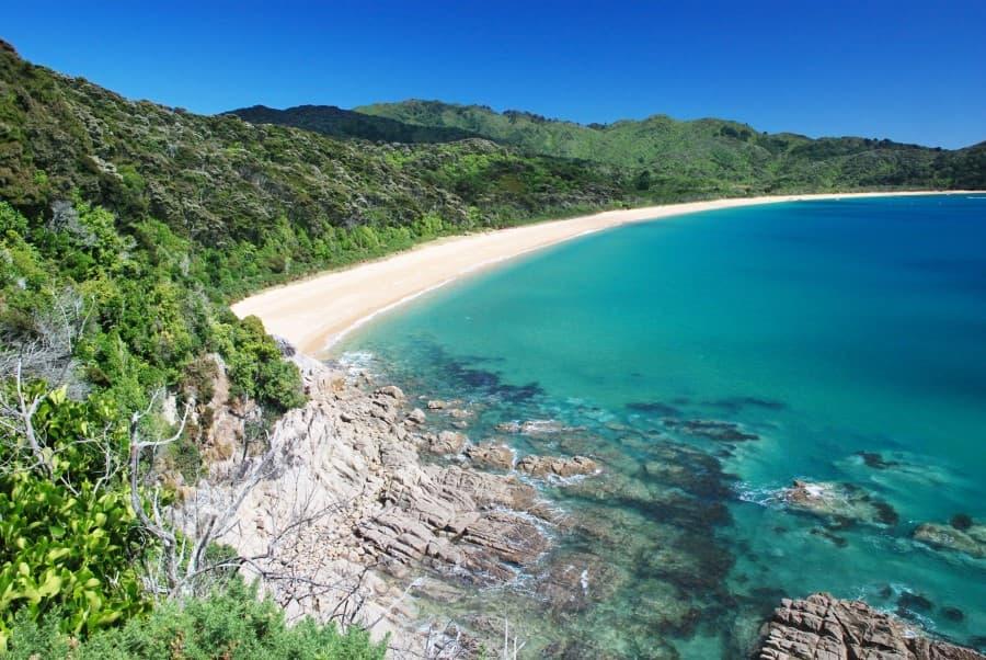A picturesque coastal landscape in Abel Tasman National Park on New Zealand's South Island, with clear turquoise water, a crescent-shaped golden sandy bay surrounded by green forested hills, and a foreground of grey rock formations that convey tranquility and seclusion.