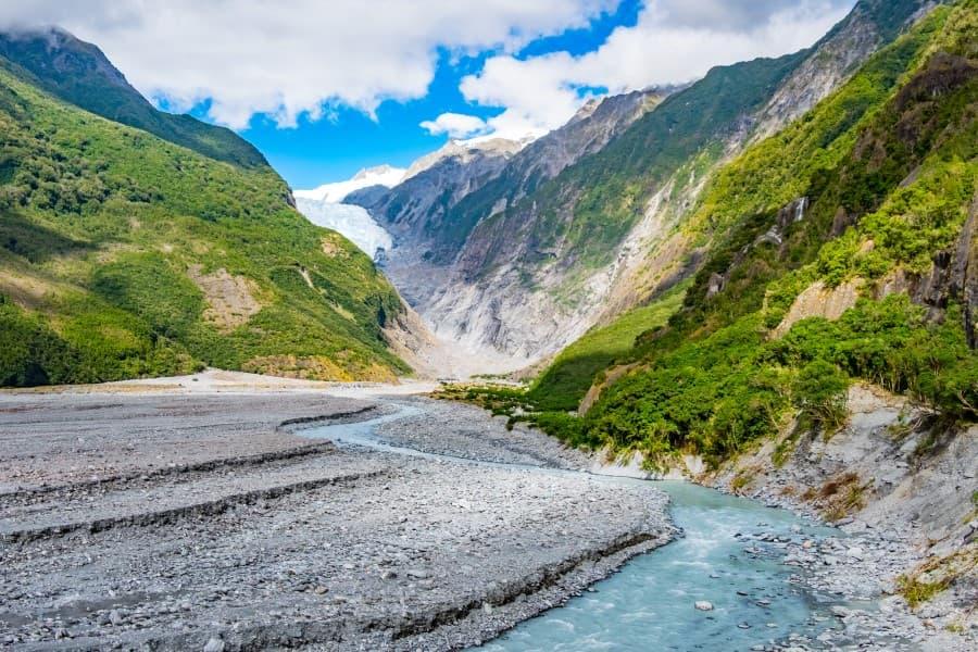 A turquoise glacial river winds through a wide stony bed, framed by lush green hills, leading the eye to the impressive ice formations of the Franz Josef Glacier, one of the famous sights on New Zealand's South Island and evoking a feeling of grandeur and untouched nature.