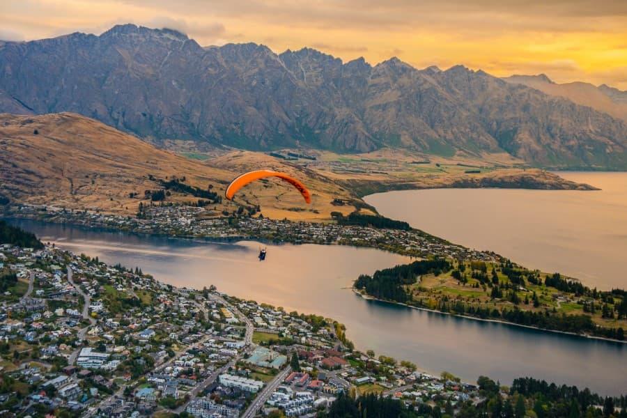 Above the picturesque town by the side of a crystal clear lake, an orange paraglider floats against the backdrop of rugged mountains, highlighting the diversity and beauty of the sights on New Zealand's South Island and conveying a sense of adventure and freedom.