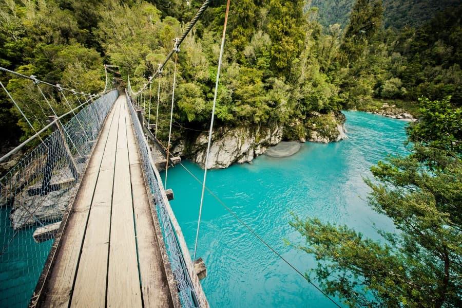 A suspension bridge stretches over the turquoise, clear waters of Hokitika Gorge, surrounded by lush greenery, a breathtaking and peaceful sight on New Zealand's South Island.