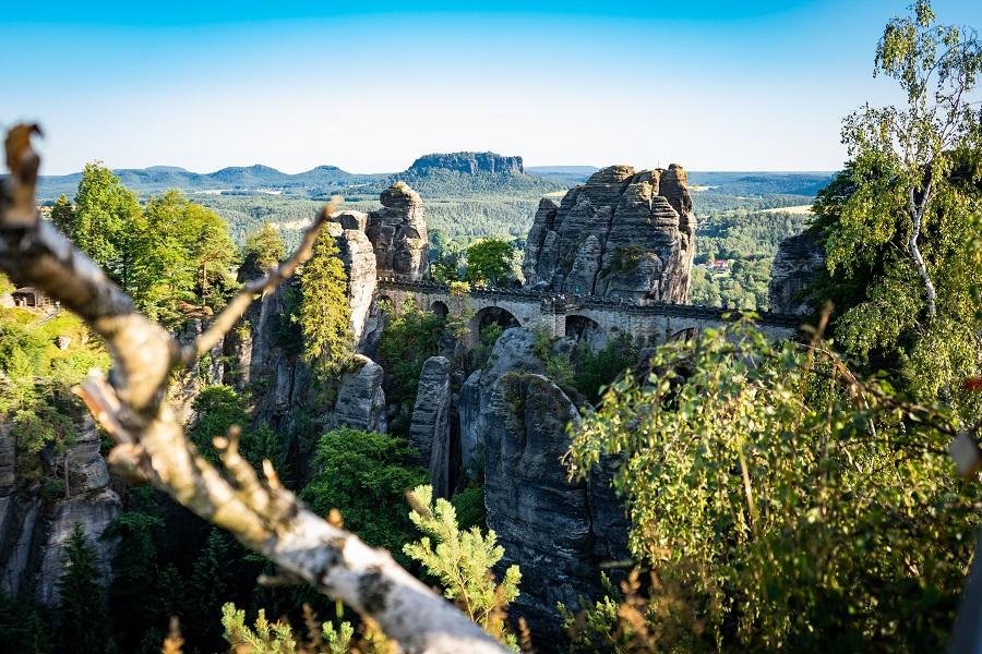The most beautiful place in Germany: Saxon Switzerland. From viewpoints like this one, where this photo was taken, you have an incredible view of the expanses of Saxon Switzerland. Green forests and trees cover the vast area and pointed rocky cliffs rise into the air repeatedly. A high bridge runs through the middle of the image.