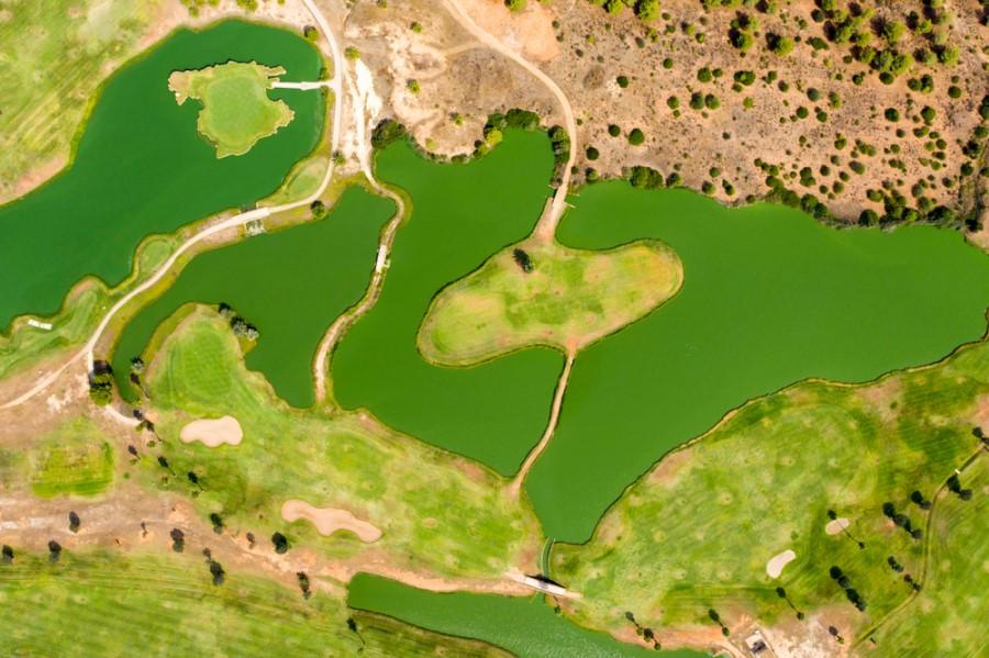 Aerial view of a golf course with several green water ponds surrounded by grass areas and sand bunkers. Paths wind through the facility. In the upper right, drought-resistant vegetation.