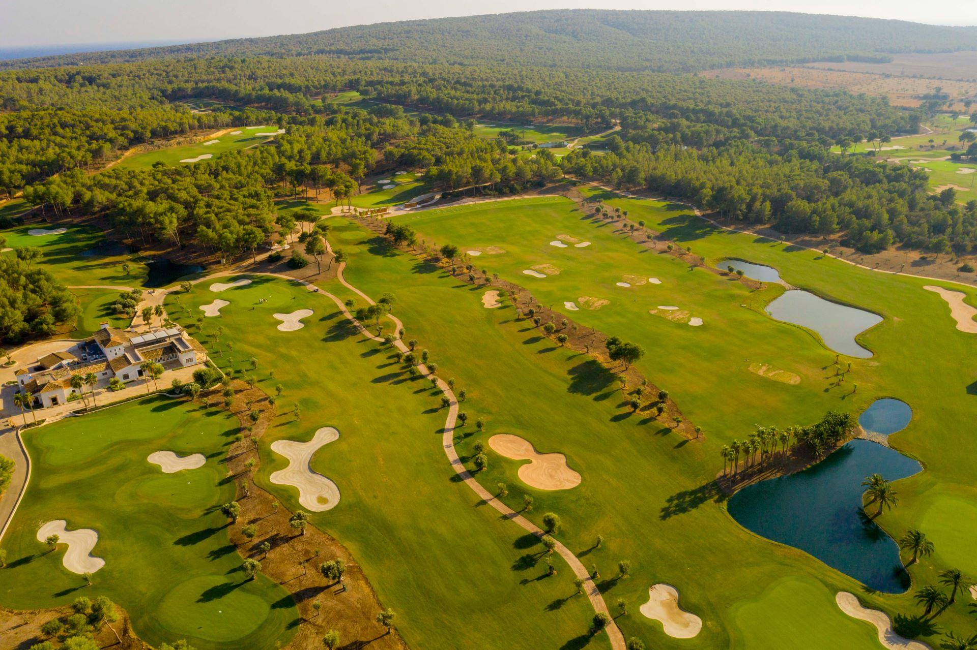 Green golf course landscape in Mallorca with flat, curved sand bunkers and ponds. In the foreground, a clubhouse; in the background, wooded hills; all at sunset.