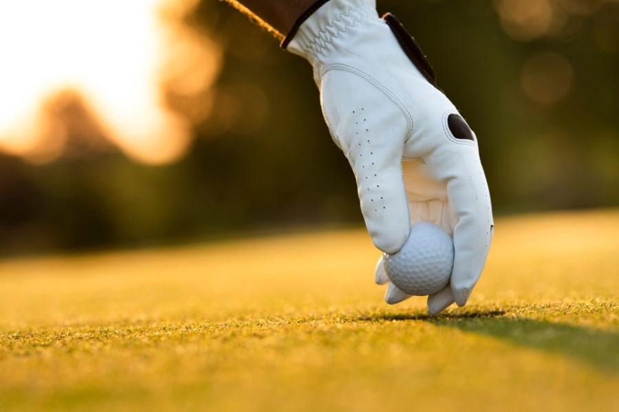 A hand with a white golf glove places a golf ball on short grass. In the background, blurred greenery in the light of the setting sun.