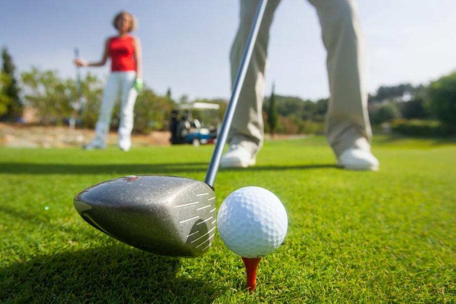 Close-up of a golf club just before a tee-off. The white golf ball sits on a red tee. In the background, a blurred person and a golf cart on green grass.