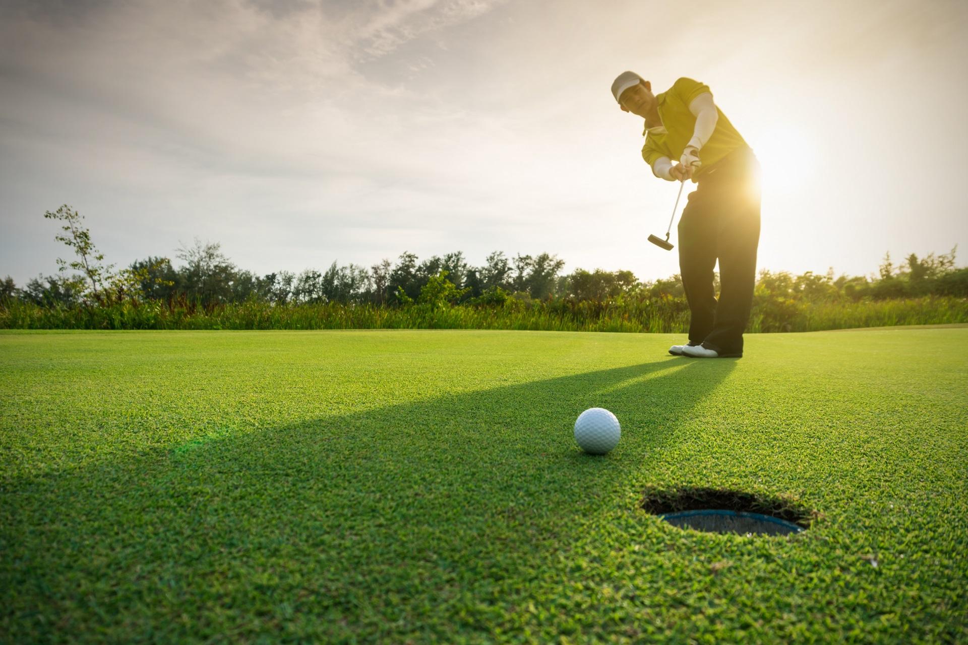 Golfer in a yellow shirt swings on green grass near a hole, golf ball nearby. Sunlight from behind, trees and shrubs in the background.