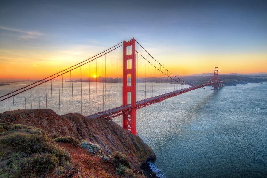 A red suspension bridge before sunset, centrally placed, stretches over blue water. The left side shows a rocky hill, the sky is colored orange and blue.
