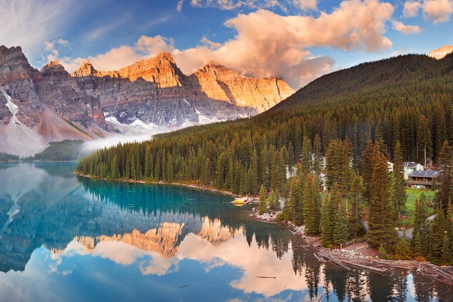 A picturesque mountain lake with turquoise water, surrounded by dense forests. In the background, sunlit mountains rise into the sky, with clouds floating above. A cabin stands on the right shore.