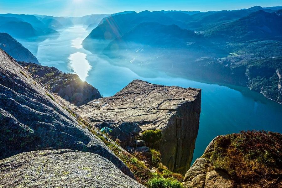 A steep cliff rises above a blue fjord. In the background, wooded mountains stretch out. The sun shines brightly, and the sky is a clear blue. Vegetation grows sporadically on the cliffs.