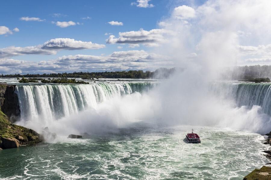 Wide waterfalls thunder down as dense mist rises. In the foreground, a red sightseeing boat sails on the river. The sky shows white clouds against a blue background.