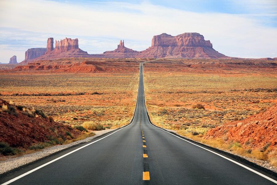 A straight asphalt road leads through a wide, red desert landscape toward rocks on the horizon, under a blue sky with white clouds.