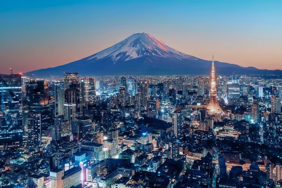 Tokyo at dusk: skyscrapers and bright lights create the skyline. The glowing Tokyo Tower is visible on the right. In the background, the snow-covered Fuji rises majestically before a colorful sky.