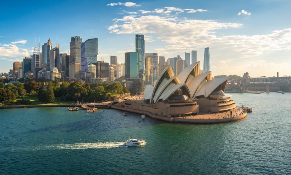 Opera House in Sydney before a skyline of skyscrapers. In the foreground, a boat crosses the blue water. Green parks lie to the left, while the sky is dotted with clouds.