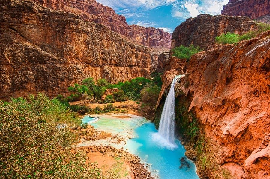 A turquoise blue waterfall cascades from red cliffs into a clear pool. Lush green vegetation surrounds the pool in the midst of a barren, rocky canyon landscape under a blue sky.
