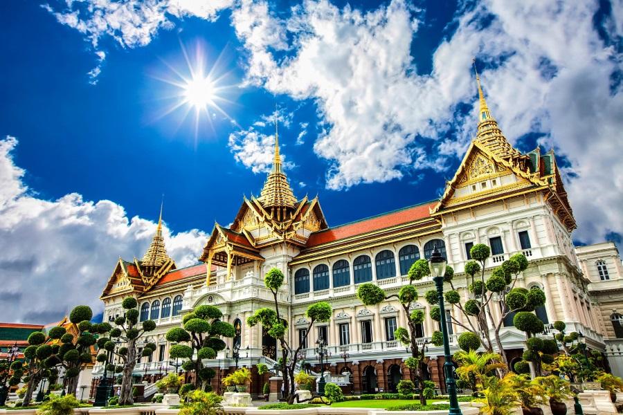 Majestic Grand Palace in Bangkok against a blue sky with white clouds, golden roofs shining in the sun. Well-kept trees and lanterns stand on green grass in front of the palace.