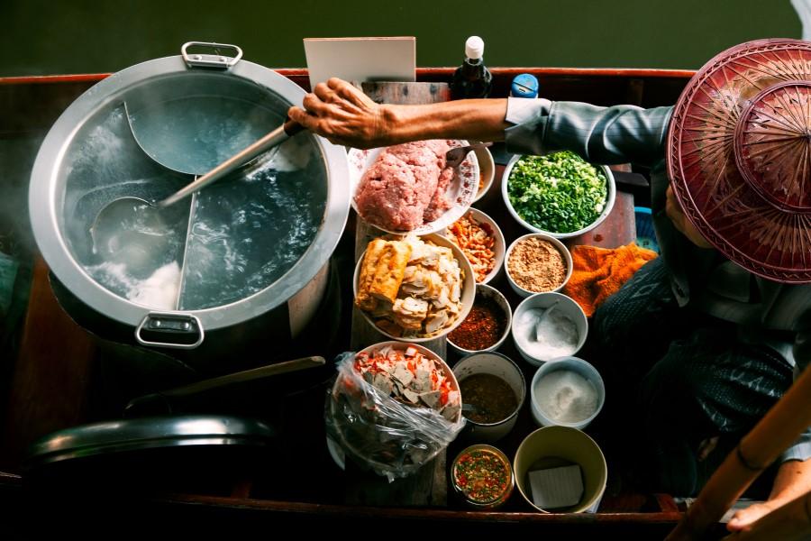 A woman sits in a boat in Bangkok, preparing fresh food in a large boiling pot of water. In front of the pot, there are many bowls of food. The woman wears a typical Thai outfit, which is decorated at the top and tapers to a point.