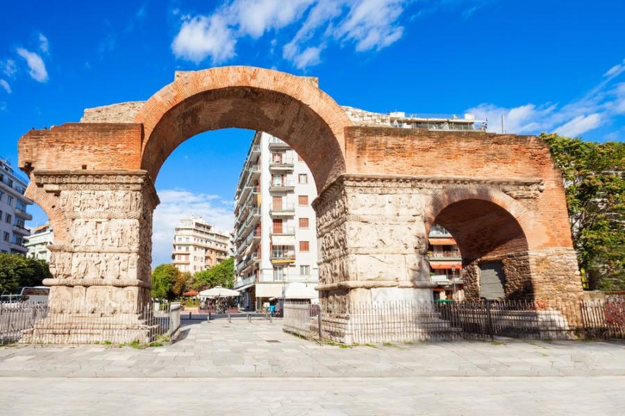 The Roman Galerius Arch decorated in the foreground, flanked by modern buildings. In the background, a clear blue sky with few clouds stretches out.