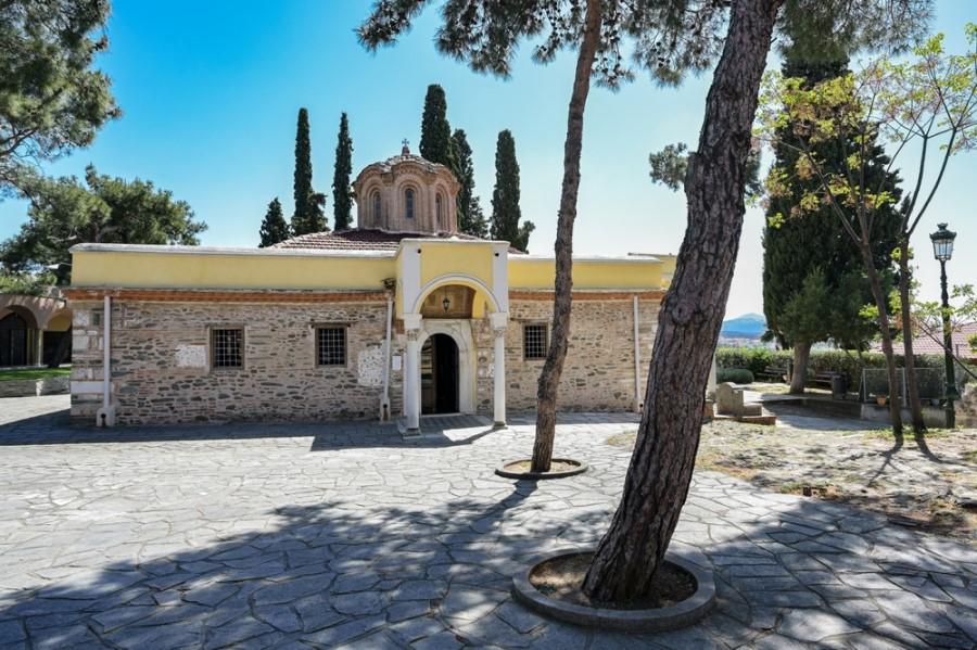 Historical monastery with yellow facade and arched doorway, surrounded by tall cypress trees. In the foreground, shade-giving trees flank a paved square.