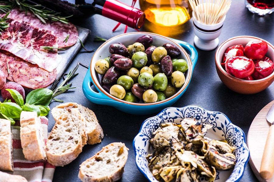 Various Spanish Tapas on a table: green and black olives in the center, stuffed peppers on the right, bread on the left, topped with sliced salami and rosemary, with artichokes in the foreground.