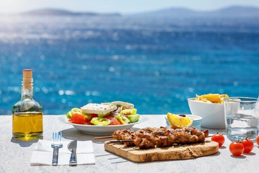Greek dishes on a table by the sea: A plate of salad and feta on the left, skewers on a wooden board in front, fries in a bowl on the right. An oil bottle, a glass of water, and tomatoes decorate the scene. The sea is in the background.