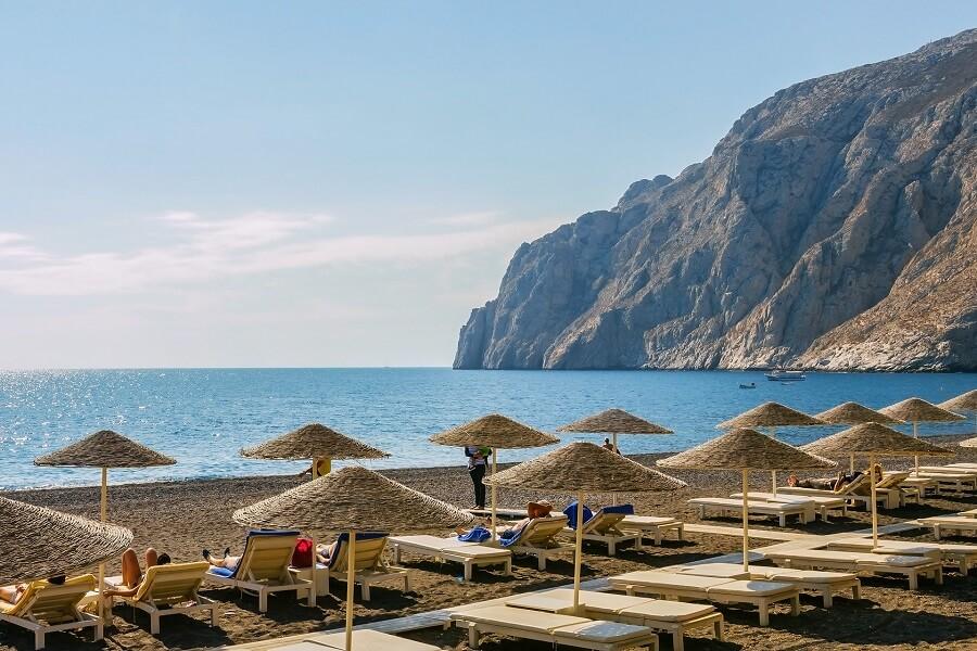Sun umbrellas and sun loungers on the black sandy beach of Kamari Beach. Crystal clear, blue sea meets a rocky cliff landscape under a clear sky.