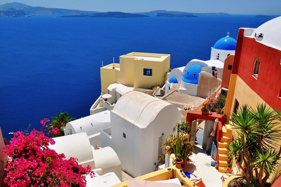 White and colorful houses with blue domes overlooking deep blue sea. Left are blooming bougainvillea, right are palm trees. The coastline stretches under a bright blue sky.