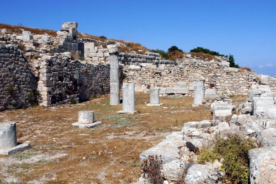 Old, light gray temple ruins with several stone columns, surrounded by grassy hills, under a clear blue sky; in the foreground loose stones and dried vegetation.