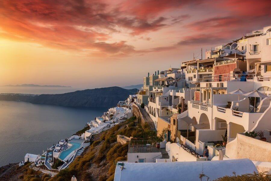 White houses with blue details cascade down a hill. In the background, the blue sea stretches under a dramatic red-orange sunset sky.