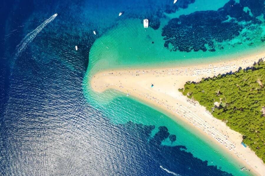 The Zlatni Rat beach on the Croatian island Brac from a bird's eye view. The majority of the image showcases the deep blue shimmering sea, which brightens and becomes more turquoise closer to the shore. The beach juts into the picture and the sea from the right half of the image. The first tip is covered with green trees, while the second tip extends further in showing the white sandy beach.