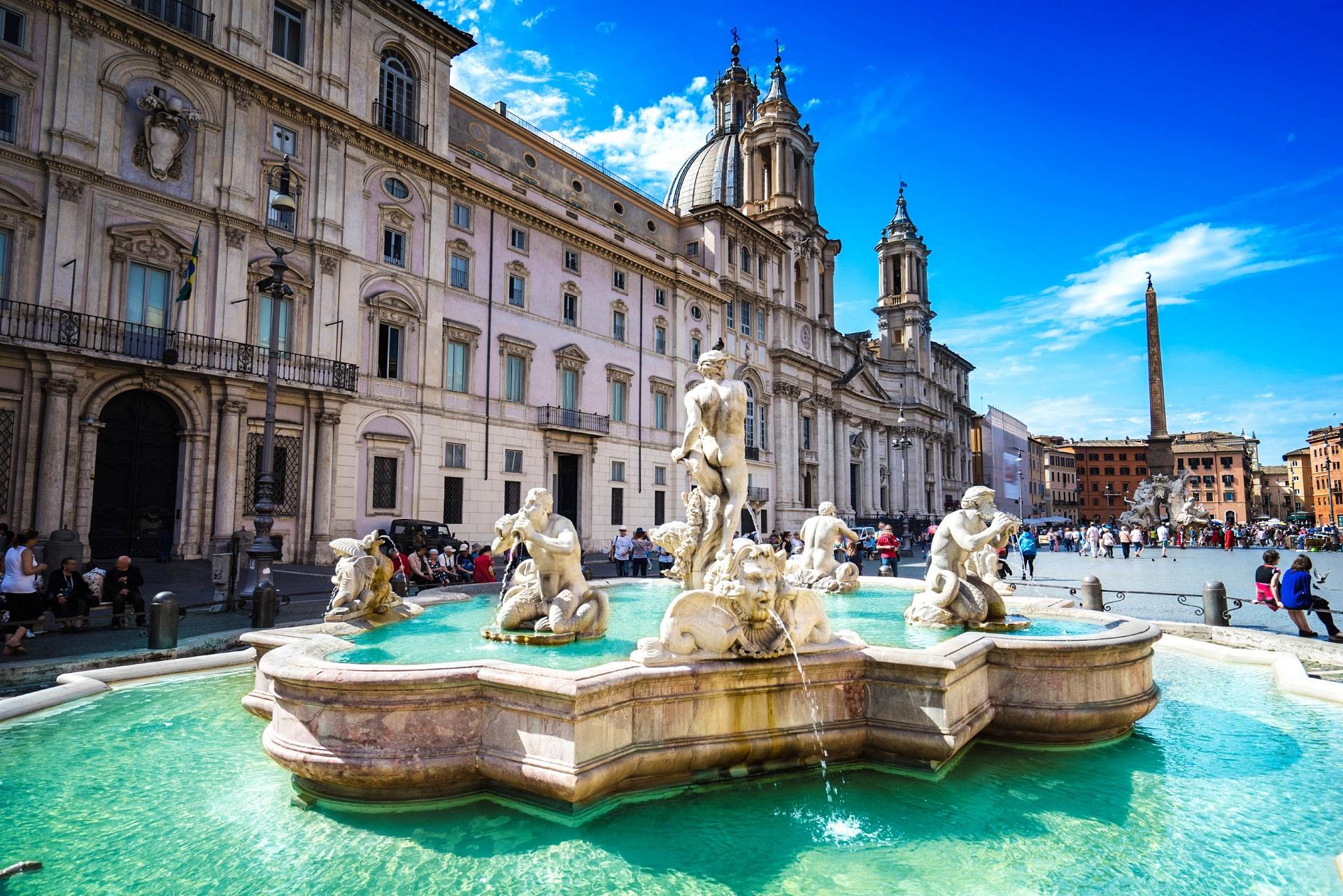 Obelisk on a fountain in the middle of Piazza Navona, surrounded by majestic baroque buildings. The sky is clear, and the cobblestones extend widely around the square.