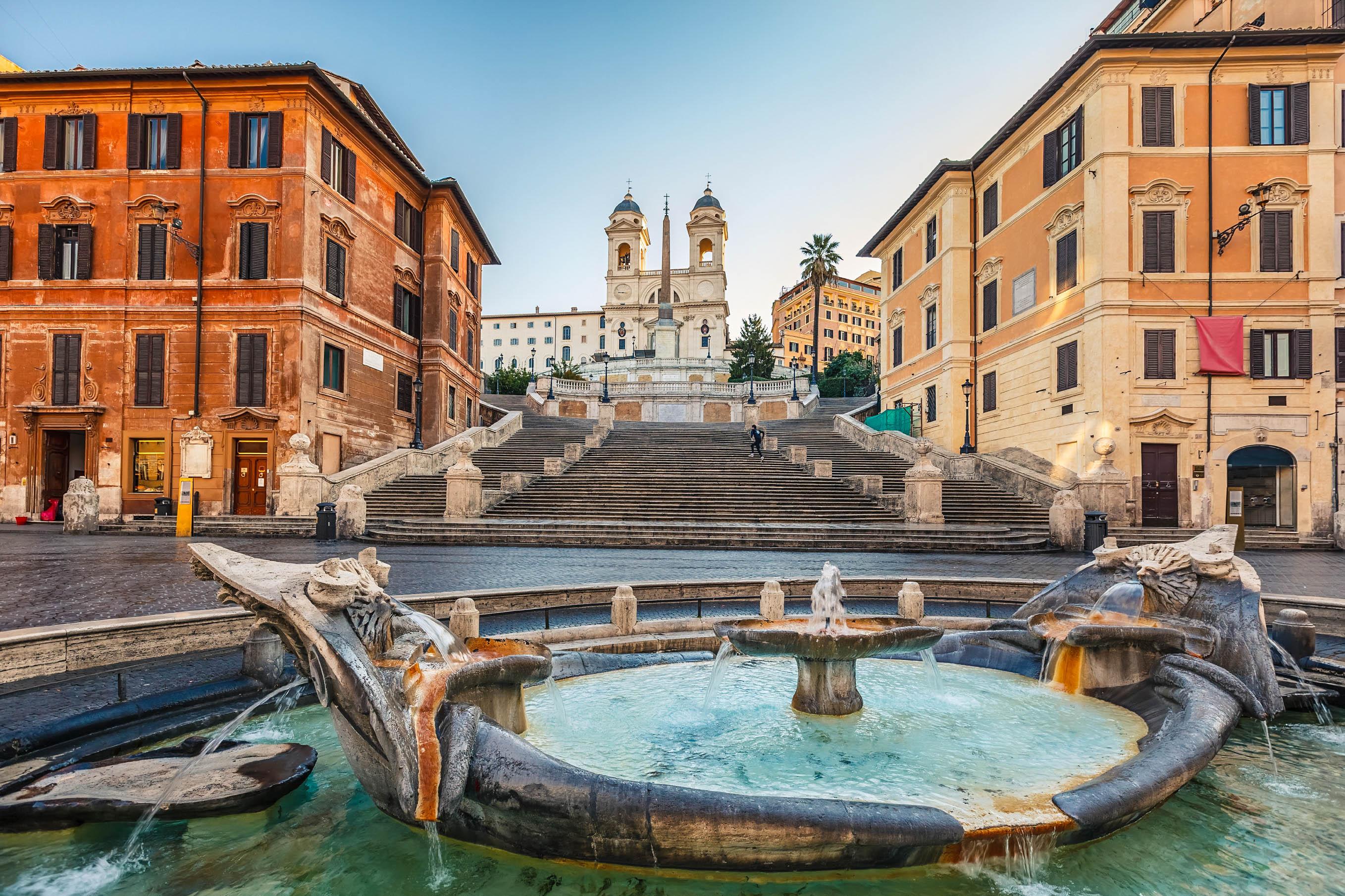 Spanish Steps in Rome at sunrise. In the foreground, a fountain, behind it the wide staircase surrounded by historic buildings. At the top, the Trinita dei Monti church stands against a clear sky.